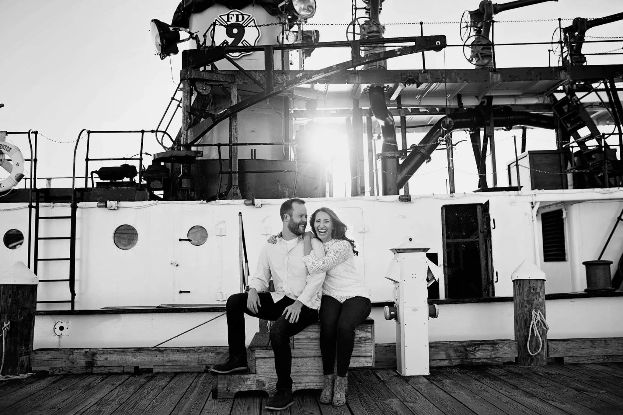 Kate and Tim sitting together on a dock in front of a large boat, smiling at each other.